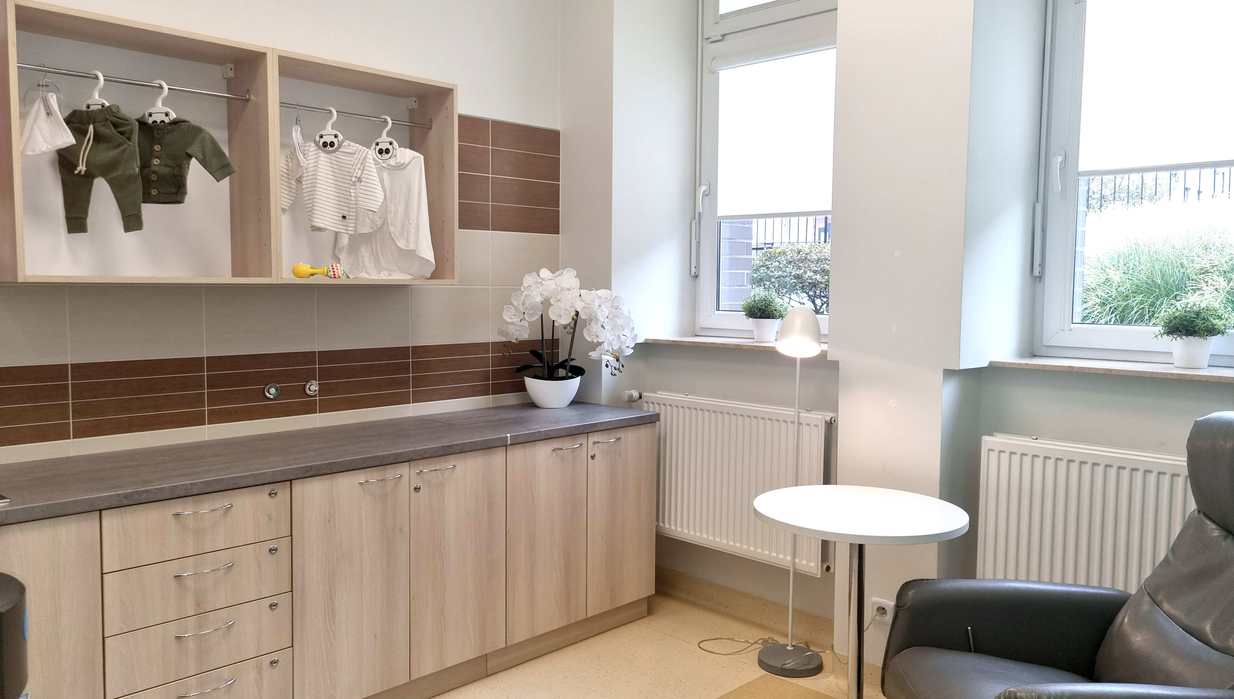 Nowoczesna kuchnia z drewnianymi szafkami, blatem, wiszącymi ubrankami dziecięcymi, oknami i fotelem, idealna do opieki nad dziećmi.